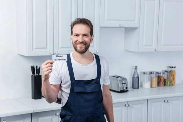 Smiling male handyman in working overall showing card with lettering home inspection — Stock Photo