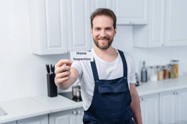 Selective focus of happy male handyman in working overall showing card with lettering home inspection — Stock Photo