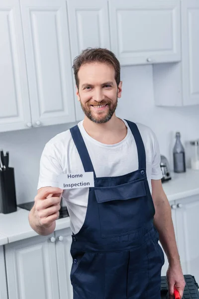 Fröhlicher männlicher Heimwerker im Arbeitsanzug zeigt Karte mit Schriftzug Home Inspection — Stockfoto