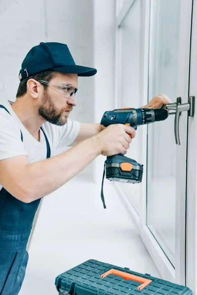 Vista lateral del reparador adulto en gafas de fijación manija de la ventana por taladro eléctrico - foto de stock