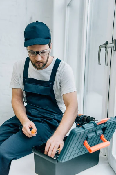 Männlicher Werkzeugmechaniker nimmt Schraubendreher aus Werkzeugkiste, während er auf Fensterbank sitzt — Stockfoto