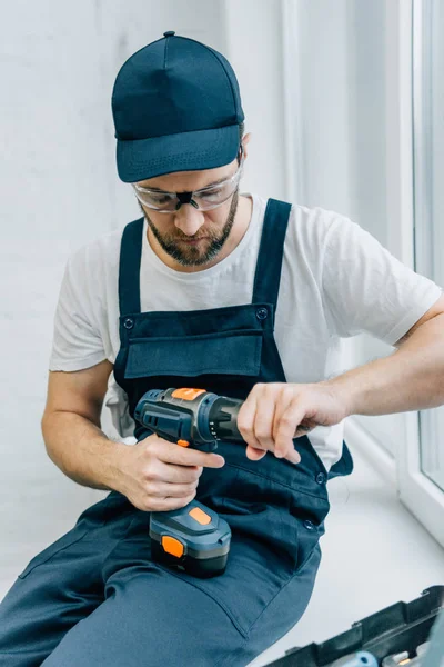 Seriöser Handwerker mit Elektrobohrmaschine sitzt auf Fensterbank — Stockfoto