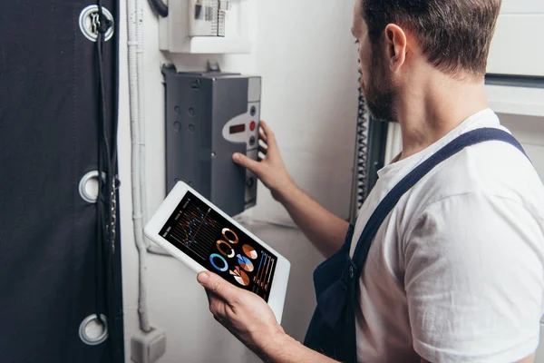 Elektriker hält digitales Tablet mit Diagrammen auf dem Bildschirm und checkt Stromkasten — Stockfoto