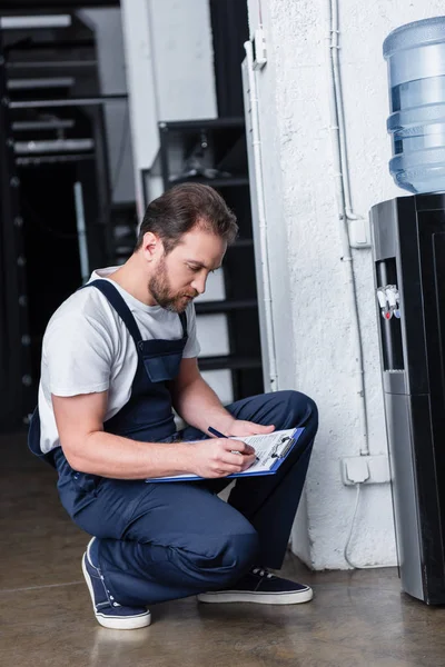 Reparador masculino escrevendo na área de transferência durante a verificação do refrigerador de água quebrado — Fotografia de Stock