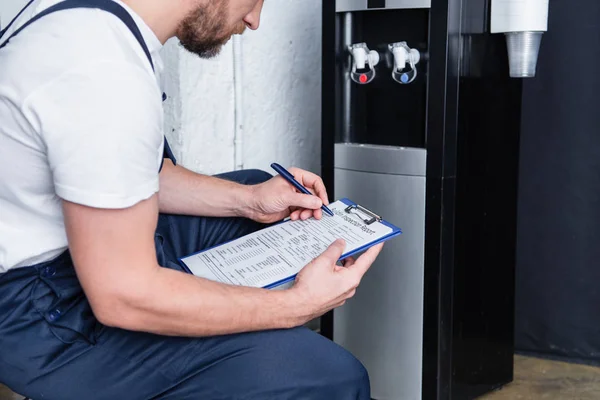 Visão parcial do reparador masculino escrevendo na área de transferência durante a verificação do refrigerador de água quebrado — Fotografia de Stock