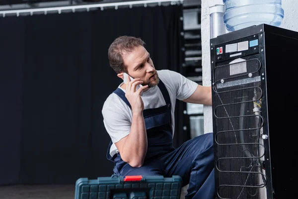 Serious male craftsman talking on smartphone during repairing water cooler — Stock Photo