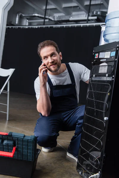 Homem adulto artesão falando no smartphone durante a reparação refrigerador de água — Fotografia de Stock