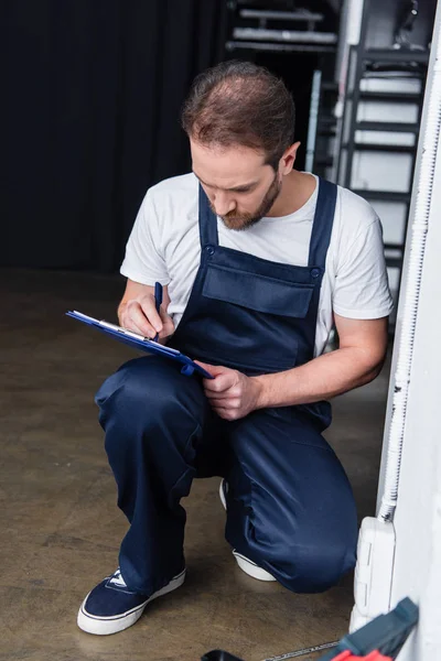 Bearded male electrician writing in clipboard near plug socket — Stock Photo