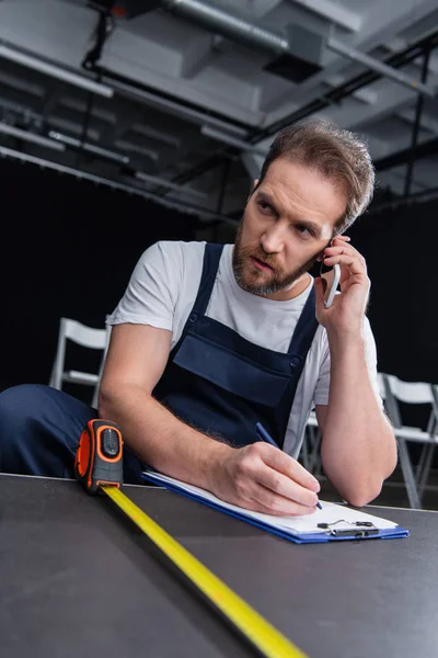 Männlicher Heimwerker spricht auf Smartphone und schreibt in Klemmbrett, während er Boden misst — Stockfoto