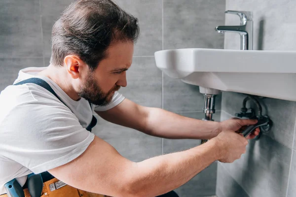 Adult male plumber in working overall fixing sink in bathroom — Stock Photo