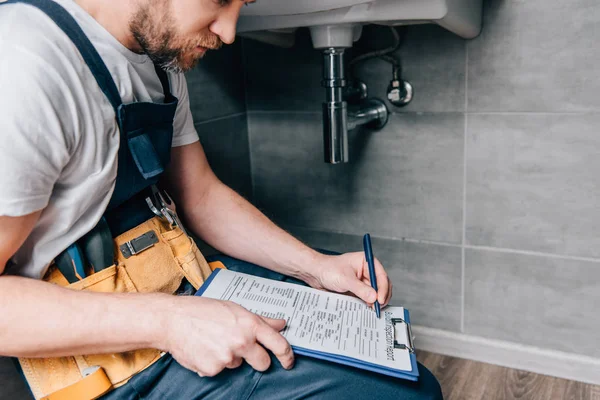 Tiro recortado de plomero masculino con la escritura del cinturón de herramientas en portapapeles cerca del fregadero roto en el baño - foto de stock