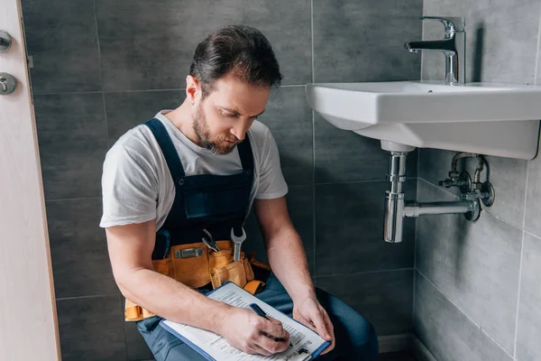 Plomero masculino adulto con la escritura de la correa de herramientas en portapapeles cerca del lavabo roto en cuarto de baño - foto de stock