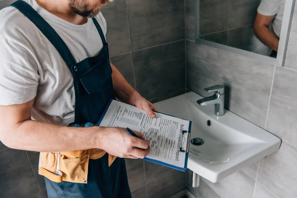 Partial view of male plumber with toolbelt writing in clipboard near broken sink in bathroom — Stock Photo