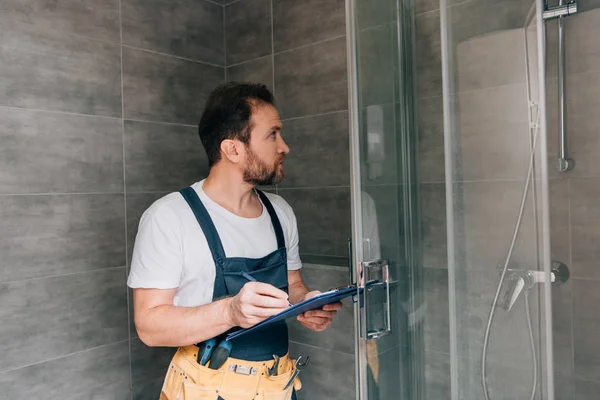 Plombier mâle adulte prenant des notes dans le presse-papiers tout en vérifiant la douche dans la salle de bain — Photo de stock