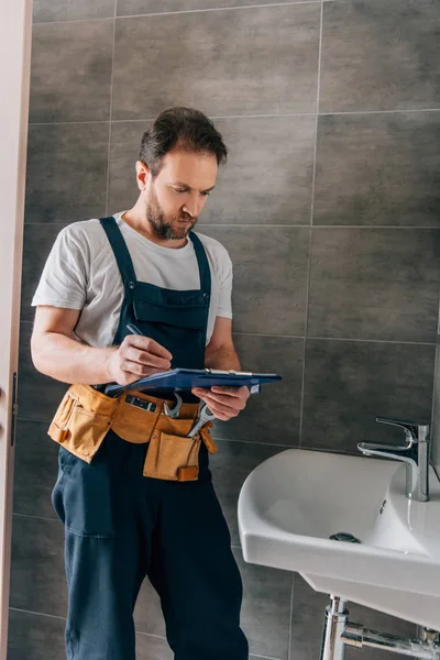 Beau plombier masculin avec ceinture d'outils écrit dans le presse-papiers près de l'évier cassé dans la salle de bain — Photo de stock