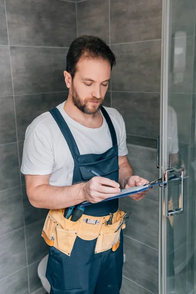 Bärtiger männlicher Klempner macht sich Notizen im Klemmbrett, während er die Dusche im Badezimmer kontrolliert — Stockfoto