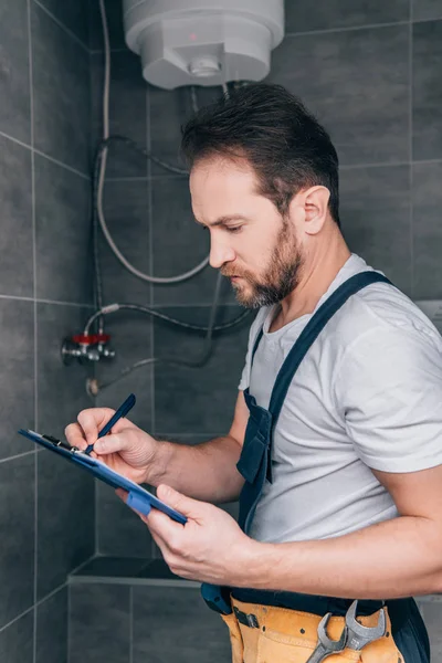 Plomero masculino adulto escribiendo en portapapeles y comprobando la caldera eléctrica en el baño - foto de stock