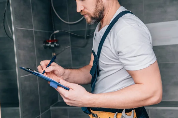 Teilansicht des Klempners, der in Klemmbrett schreibt und Elektroboiler im Badezimmer überprüft — Stockfoto