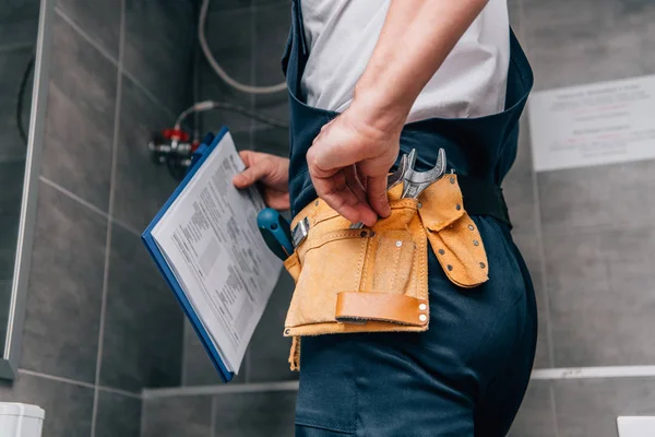 Tiro recortado de plomero masculino con portapapeles y cinturón de herramientas de pie en el baño - foto de stock