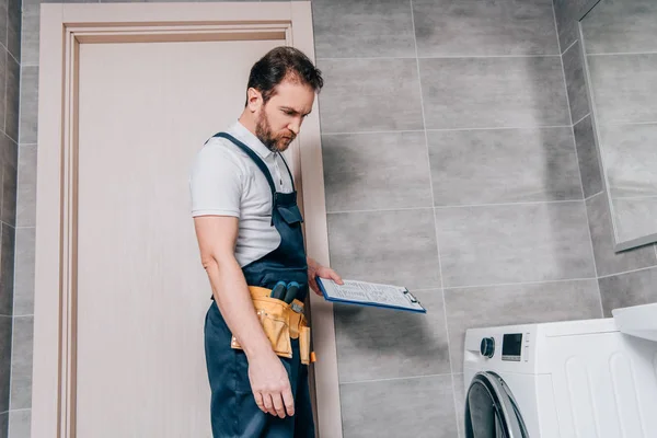 Repairman with toolbelt and clipboard checking washing machine in bathroom — Stock Photo