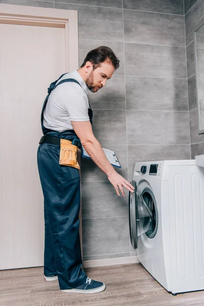 Männliche erwachsene Reparaturkraft mit Werkzeuggurt und Klemmbrett überprüft Waschmaschine im Badezimmer — Stockfoto