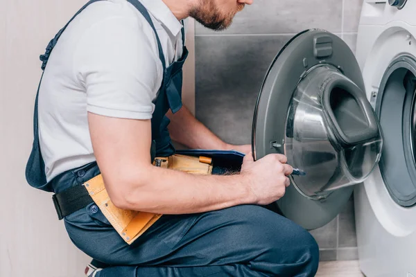 Schnappschuss eines Heimwerkers mit Werkzeuggurt und Klemmbrett, der Waschmaschine im Badezimmer kontrolliert — Stockfoto