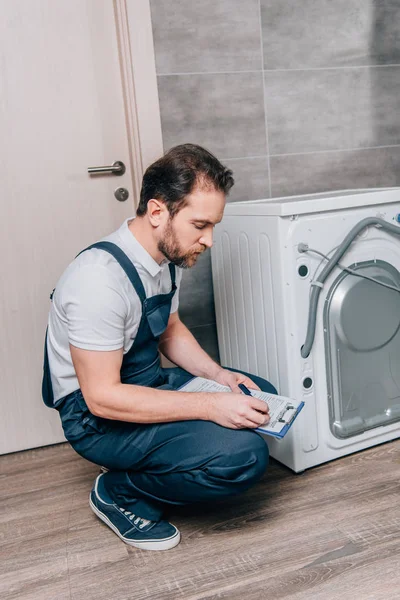 Adulto faz-tudo escrevendo na área de transferência e verificando máquina de lavar roupa no banheiro — Fotografia de Stock