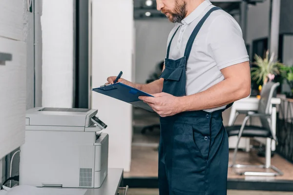 Partial view of craftsman checking copy machine and writing in clipboard in modern office — Stock Photo