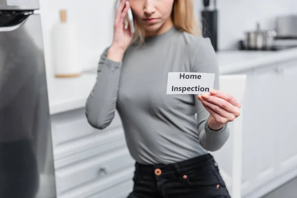 Partial view of young woman holding card with lettering home inspection and talking on smartphone in kitchen — Stock Photo