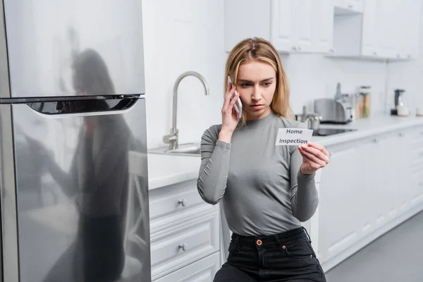 Junge Frau hält Karte mit Schriftzug nach Hause und telefoniert auf Smartphone neben kaputtem Kühlschrank in Küche — Stockfoto