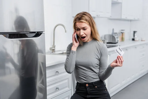 Angry young woman holding card with lettering home inspection and yelling on smartphone in kitchen near broken refrigerator — Stock Photo