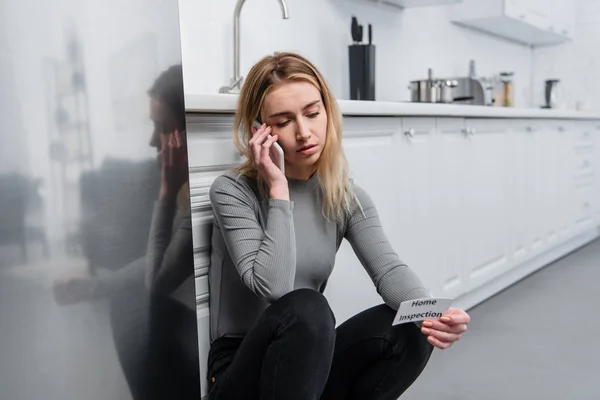 Blonde young woman holding card with lettering home inspection and talking on smartphone in kitchen near broken fridge — Stock Photo