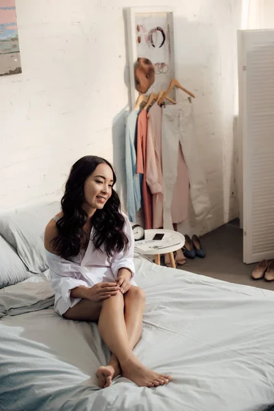 Красива усміхнена азіатська дівчина в білій сорочці сидить на ліжку і дивиться геть — стокове фото