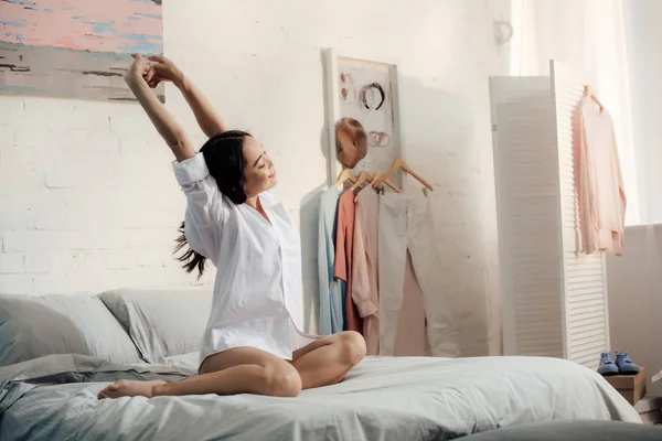 Attraente asiatico donna in bianco camicia seduta su letto e stretching mani in il mattina — Foto stock