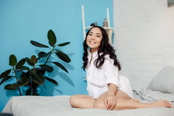 Hermosa sonriente joven asiático mujer en blanco camisa sentado en la cama en la mañana - foto de stock