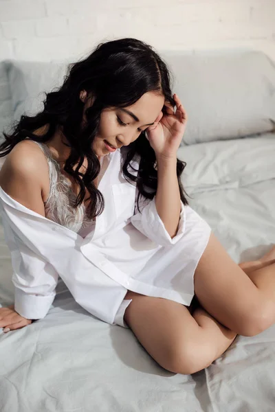 Alto ángulo vista de hermosa sonriente chica asiática en lencería y camisa blanca sentado en la cama y mirando hacia abajo - foto de stock