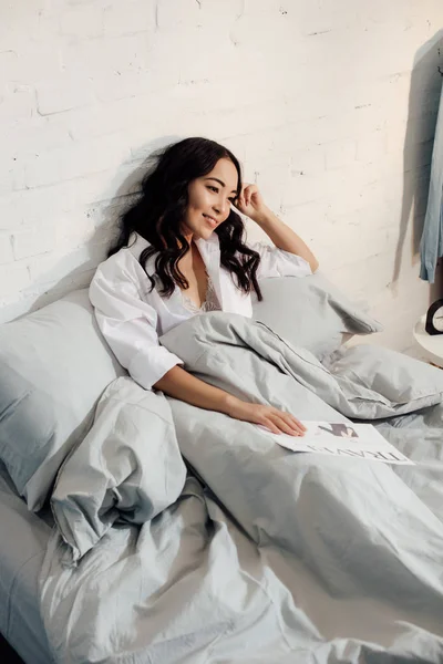 Высокий угол зрения на красивую улыбающуюся молодую женщину лежащую в постели с газетой путешествия — стоковое фото