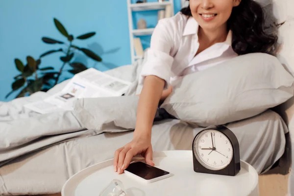 Recortado disparo de sonriente mujer joven acostada en la cama y la celebración de teléfono inteligente con pantalla en blanco - foto de stock