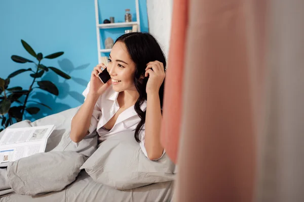 Hermosa sonriente chica asiática acostada en la cama y hablando por teléfono inteligente - foto de stock