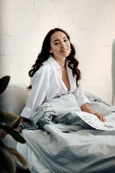 Attractive asian woman holding travel newspaper and smiling at camera while sitting on bed — Stock Photo