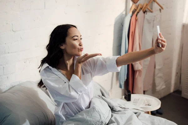 Attractive asian woman blowing kiss and taking selfie with smartphone in bed — Stock Photo