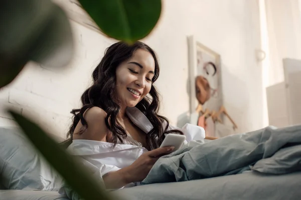 Baixo ângulo vista de belo sorriso asiático menina usando smartphone na cama — Fotografia de Stock