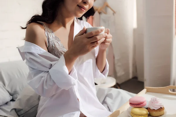 Cropped shot of smiling asian girl in bra and white shirt holding cup of coffee and sitting on bed — Stock Photo
