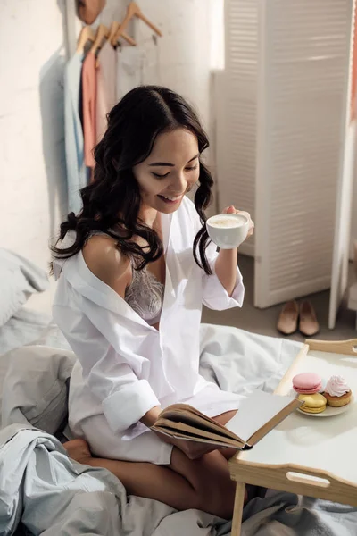 Schöne lächelnde asiatische Mädchen Buch lesen und Kaffee trinken auf Bett — Stockfoto