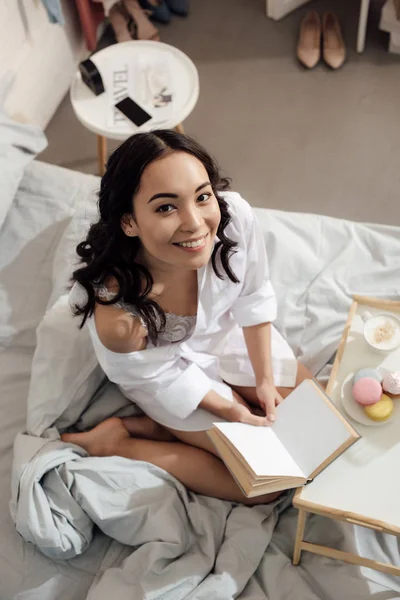 Vue grand angle de belle fille asiatique heureuse assis avec livre sur le lit et souriant à la caméra — Photo de stock