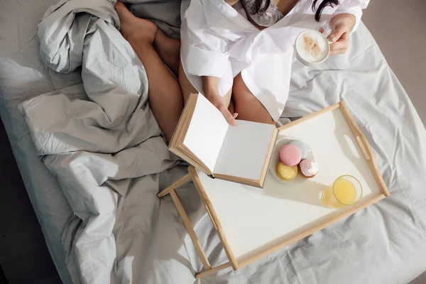 Schnappschuss einer jungen Frau in Dessous und Hemd, die eine Tasse Cappuccino in der Hand hält und Buch auf dem Bett liest — Stockfoto