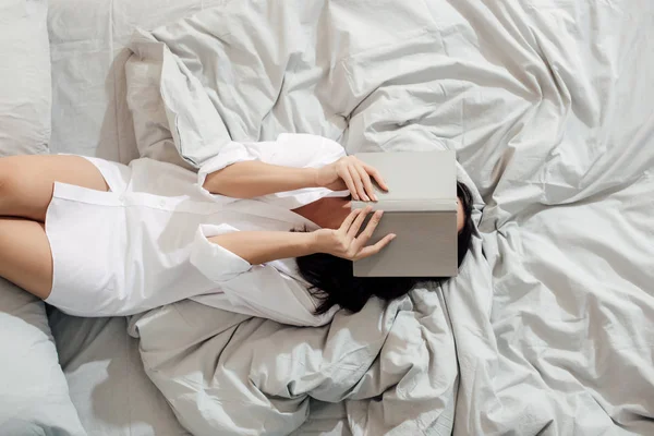 Top view of young woman in white shirt lying with book on face in bed — стоковое фото