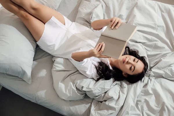 Вид зверху красива усміхнена азіатська дівчина читає книгу в ліжку — стокове фото