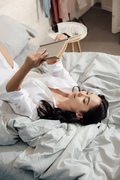 Alto ângulo vista de atraente asiático menina leitura livro no cama — Fotografia de Stock