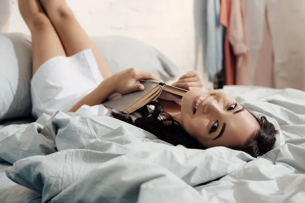 Happy young asian woman lying on bed with book and smiling at camera — Stock Photo